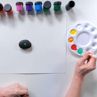 Person painting a rock with a paintbrush next to a palette of colors on a white surface.