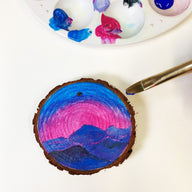 Painted wooden slice with a mountain landscape and a paintbrush next to a palette of colors.